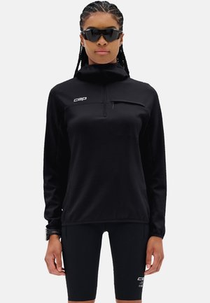 Vrouw draagt een zwarte hooggesloten top met lange mouwen en rits en zwarte fietsshorts, met een zonnebril en polshorloge op een witte achtergrond.