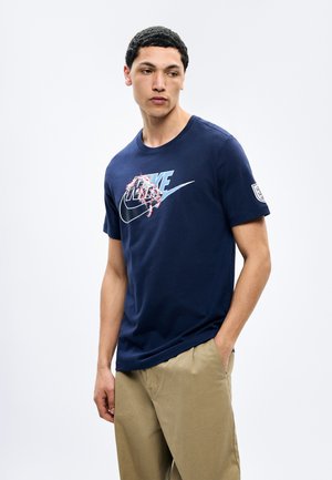 Jeune homme aux cheveux courts et bouclés portant un t-shirt Nike bleu marine avec un logo graphique et un pantalon beige, debout avec une main dans la poche.