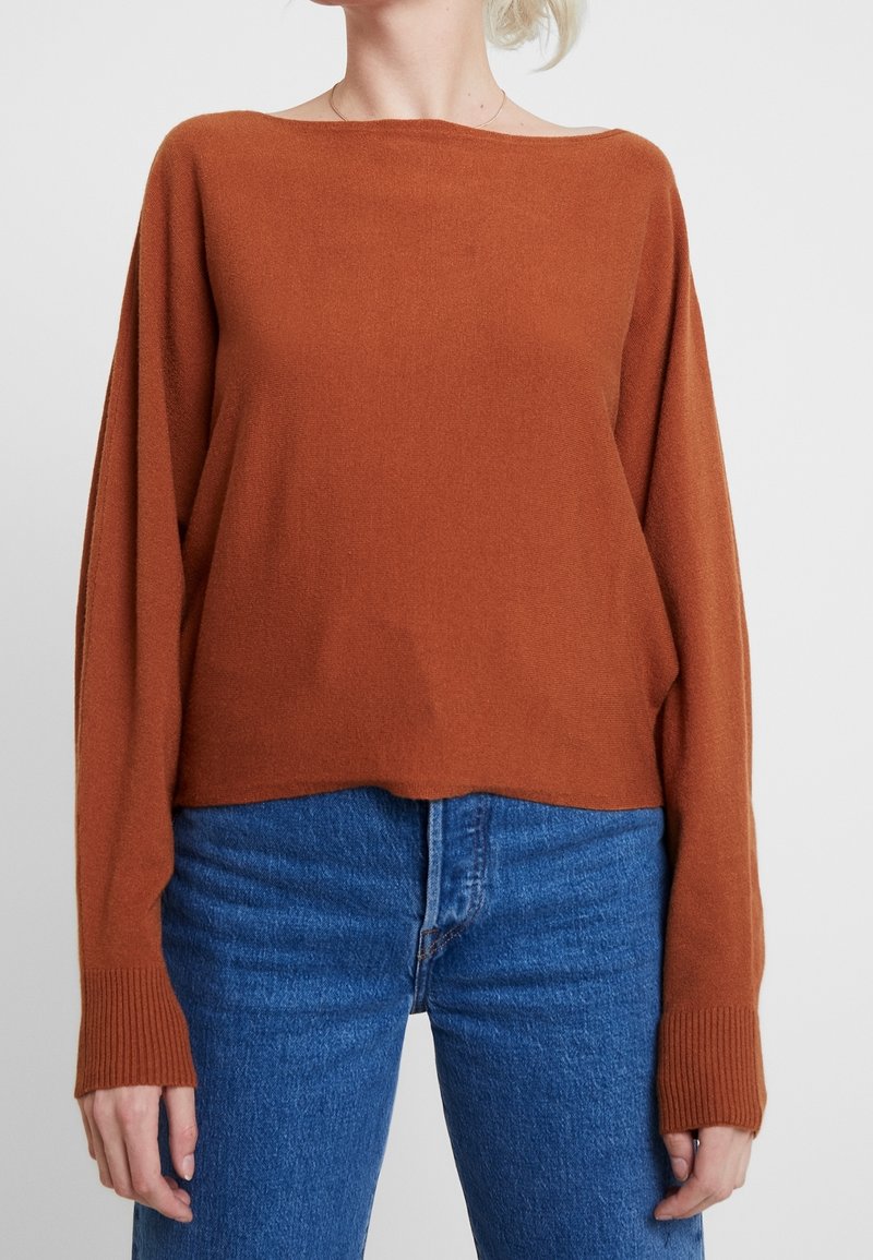 Personne portant un pull à manches longues couleur rouille avec des poignets côtelés et un jean bleu taille haute sur un fond uni.