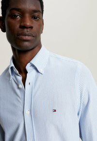 Camisa de botones a rayas azul claro con cuello puntiagudo. Presenta un pequeño logo en el pecho y botones blancos. Textura de tela suave.