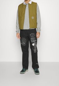 Olivgrön vadderad väst över grå långärmad tröja; svarta byxor med vita grafiska tryck; gröna och svarta sneakers.