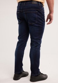 Muškarac obučen u tamnoplave uske traperice i crne tenisice, stoji raširenih nogu, vidljiv od struka do stopala, na svijetloj pozadini.