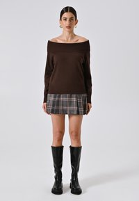 Brauner schulterfreier Pullover mit einem karierten Minirock in Blau- und Beigetönen, kombiniert mit schwarzen knielangen Stiefeln. Glatte Textur.