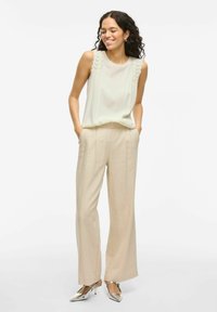 Top sin mangas de crema claro con detalles de encaje en los hombros, combinado con pantalones de pierna ancha beige y zapatos metálicos de punta. 