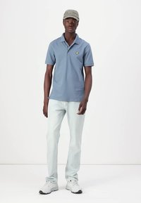 Lyseblå polo med korte ermer og en gul logo, parret med lyse jeans og hvite sneakers. Avslappet antrekk med en nøytral caps.