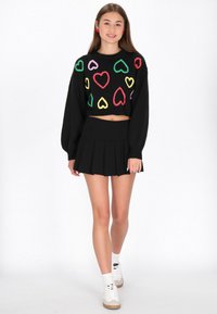 Fekete cropped pulóver színes szívmintákkal, fekete rakott szoknyával párosítva. A modellen fehér sneaker cipők és bokazoknik vannak.