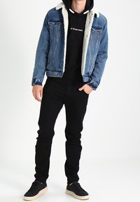 Denim jacket with a shearling collar, blue color, front pockets, worn over a black hoodie and paired with black pants and black shoes.