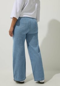 Henkilö, joka on pukeutunut vaaleansinisiin löysiin farkkuihin ja tilavaan valkoiseen paitaan, seisoo selkä kameraan päin yhtenäistä taustaa vasten.