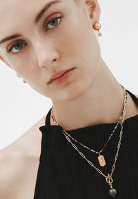 Collares de oro y cuentas en capas sobre una blusa negra, con un colgante de diseño de destello solar y una piedra preciosa verde. Pequeños pendientes de botón de oro.