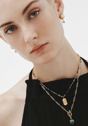 Collares de oro y cuentas en capas sobre una blusa negra, con un colgante de diseño de destello solar y una piedra preciosa verde. Pequeños pendientes de botón de oro.