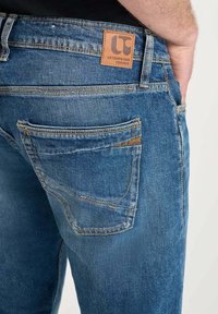 Jeans de mezclilla en azul con una etiqueta de cuero. Las características incluyen un diseño de corte recto y dos bolsillos traseros con costura en contraste.