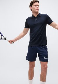 Νέος άνδρας με μπλε αθλητική μπλούζα και σορτς κρατάει ρακέτα τένις, έτοιμος να χτυπήσει, μπροστά σε λευκό φόντο.