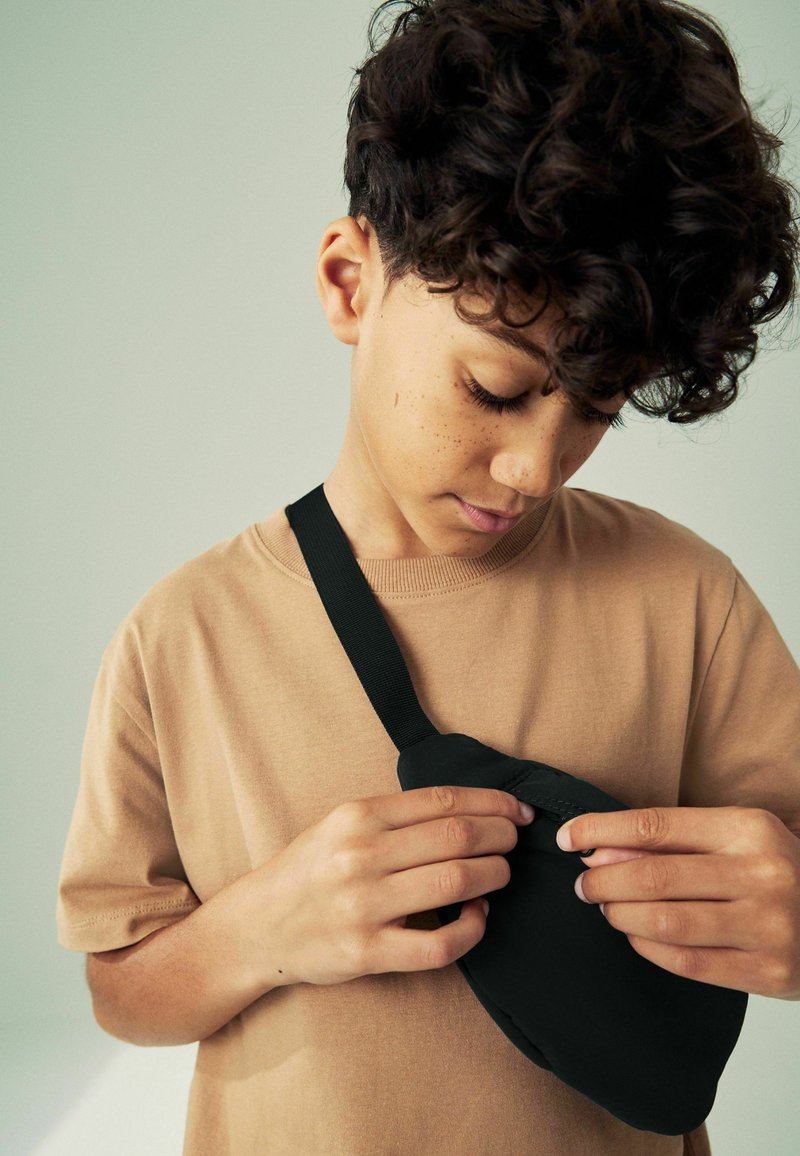 Sac bandoulière noir avec une texture lisse et une sangle réglable, tenu par une personne portant un t-shirt beige, focalisé sur la fermeture du sac.