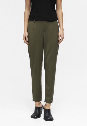 Femme portant un pantalon sur-mesure longueur cheville couleur vert olive, un top noir sans manches et des mules noires à talons, debout devant un fond blanc.