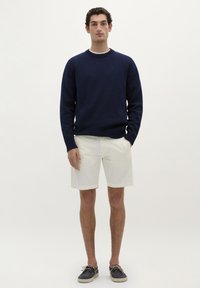 Marine strikketrøje, hvide shorts og marine bådsko. Trøjen har en ribbet halsudskæring og mansjetter. Shortsene er knælange og ensfarvede.