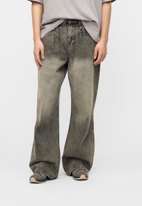 Άτομο με φαρδιά, ξεθωριασμένα μαύρα τζιν παντελόνια και γκρι αθλητικά παπούτσια, στέκεται μπροστά σε απλό λευκό φόντο με τα χέρια χαλαρά δίπλα στο σώμα.