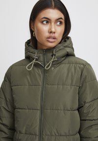 Olive zaļš pūkains mētelis ar rāvējslēdzēju priekšpusē, regulējamu kapuci un quilted tekstūru. Ievērojami ir uzgriežņi un tīrs, vienkāršs dizains.