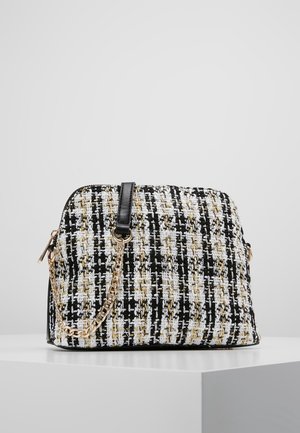 Musta, valkoinen ja kulta tweed-käsilaukku kultaisella ketjuolkaimella ja mustilla nahkakoristeilla, esillä valkoisella jalustalla.