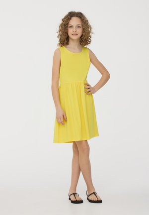 Vestito giallo senza maniche realizzato in tessuto leggero, con un corpo aderente e una gonna svasata. Indossato con sandali neri, esposto su uno sfondo bianco.