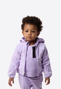 Lilla fleece hettegenser med glidelås, hette, frontlomme og svart panel. Matcher med et ensfarget lilla antrekk, med enkelt design og teksturert stoff.