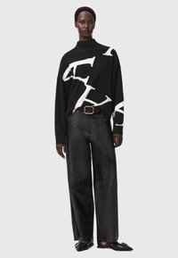 Czarny sweter dzianinowy z białymi abstrakcyjnymi literami, wysoki kołnierz i luźny fason; w połączeniu z czarnymi skórzanymi spodniami o szerokiej nogawce i czarnymi butami.