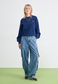 Marineblå hettegenser med rødt hjerte-logo, kombinert med løse lyseblå cargo-jeans med sidelommer. Modellen har på seg mørke sko.