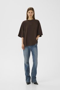 Young woman wearing oversized dark brown t-shirt, blue straight-leg jeans, and black pointed shoes, standing against white background.