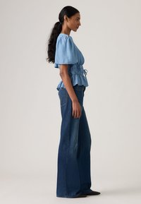 Ανοιχτό μπλε βαμβακερό πουκάμισο με κοντά φουσκωτά μανίκια, σφιχτή μέση και βολάν στο τελείωμα, συνδυασμένο με ψηλόμεσα σκουρόχρωμα μπλε τζιν καμπάνα.