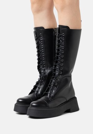 Bottes noires montantes en cuir brillant avec un laçage à l'avant, bout rond et une semelle épaisse. Fermeture éclair sur le côté pour un enfilage facile.