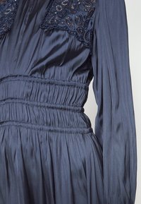 Robe en satin bleu foncé avec manches longues, taille froncée et détails en dentelle brodée près de l'encolure.