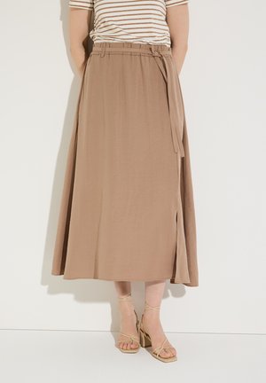 Frau trägt einen beigen A-Linien-Midirock mit seitlichem Schlitz und gebundenem Gürtel, kombiniert mit beigen Sandalen mit Blockabsatz und Riemen.