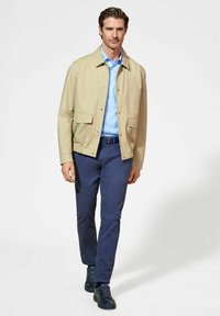 Beżowa lekka kurtka z dwoma przednimi kieszeniami na jasnoniebieską koszulę, w połączeniu z ciemnoniebieskimi spodniami i czarnymi sznurowanymi butami.