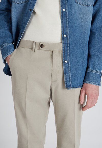 Denimjack in medium blauw over een crème trui, gecombineerd met lichtbeige plooibroek met een knop aan de voorkant en zijzakken.