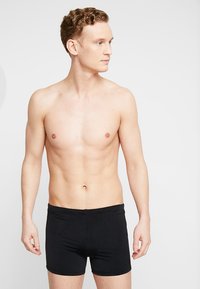 Fekete boxeralsók, puha anyagból készült, szoros szabásúak és sima derékpánttal rendelkeznek. A modell egyenesen áll, semleges testtartással és arckifejezéssel.