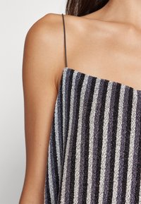 Vrouw draagt een mouwloze top met verticale strepen in zwart, grijs en wit, voorzien van dunne zwarte spaghetti-bandjes.