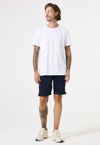 Weißes Baumwoll-T-Shirt, marineblaue Shorts mit umgeschlagenen Säumen und beige Sneaker. Sichtbare Tattoos an Armen und Beinen. Schlichter, lässiger Stil.