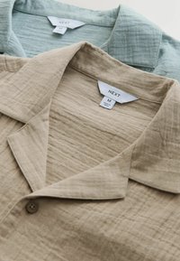 Két közepes méretű, texturált gombos ing, az egyik bézs, a másik világoszöld, "NEXT" márkajelzésű címkékkel és hegyes gallérokkal.