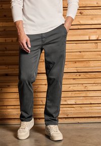 Harmaat chinot, valmistettu puuvillasekoitteesta, joissa on slim fit -leikkaus ja sivutaskut, yhdistetty valkoisiin lenkkareihin. Taustalla puupanelointi.