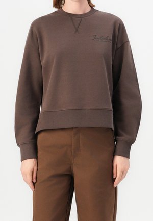 Osoba ubrana w brązową bluzę Timberland i pasujące brązowe spodnie, stojąca na jednolitym białym tle.