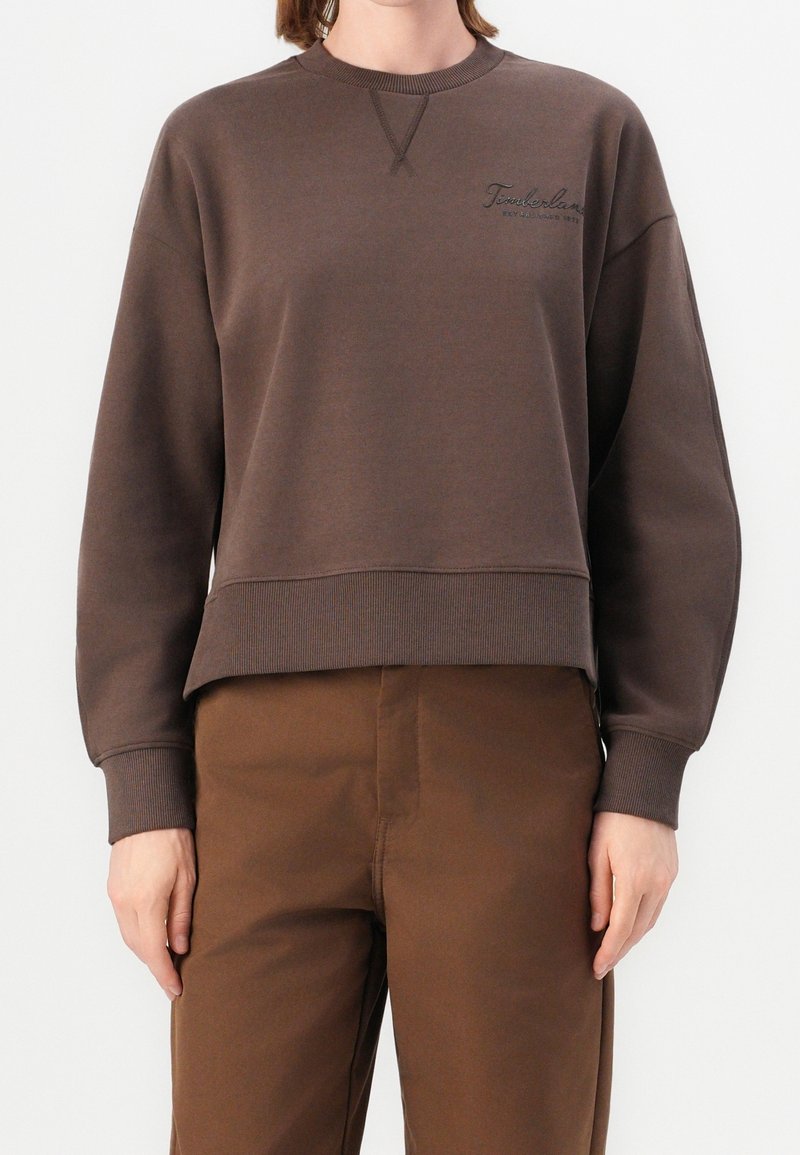 Personne portant un sweat Timberland marron et un pantalon marron assorti, debout devant un fond blanc uni.