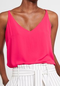 Haut rose sans manches avec un décolleté en V, fabriqué en tissu léger, présentant une coupe décontractée et une texture lisse. Pantalon rayé taille haute visible.