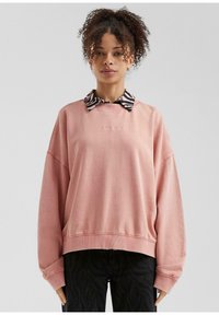 Sudadera rosa de gran tamaño con puños acanalados, superpuesta a un cuello a rayas en blanco y negro. Texto "La Clínica QS Low" bordado en la parte delantera.