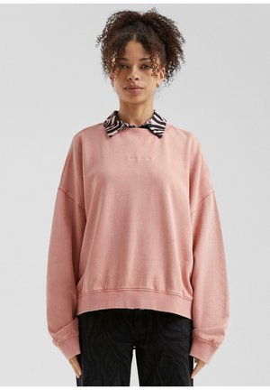 Sweatshirt surdimensionné rose avec des poignets côtelés, superposé sur un col rayé noir et blanc. Texte "The QS Low Clinic" brodé à l'avant.