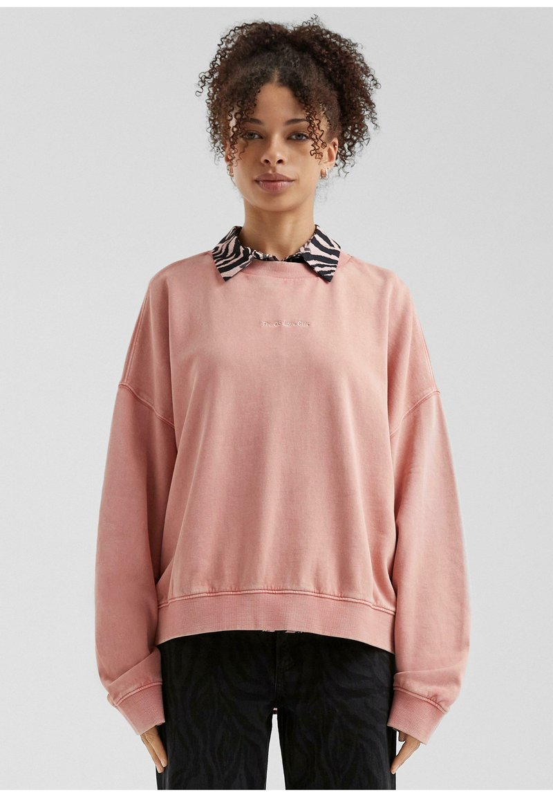 Sudadera rosa de gran tamaño con puños acanalados, superpuesta a un cuello a rayas en blanco y negro. Texto "La Clínica QS Low" bordado en la parte delantera.
