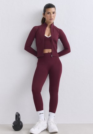 Frau in bordeauxrotem Sportoutfit mit weißen Sneakers und Socken, die an einer Wand neben einer schwarzen Kettlebell auf dem Boden steht.