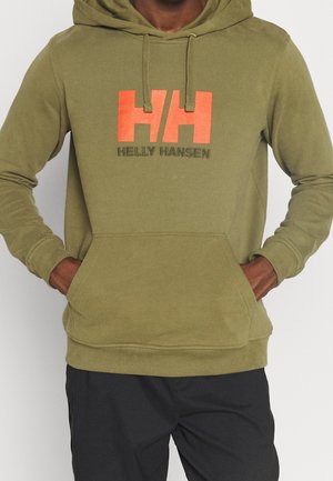 Person trägt olivgrünen Hoodie mit orangefarbenem "HH"- und schwarzem "Helly Hansen"-Logo, Hände in der Fronttasche, kombiniert mit schwarzen Hosen.