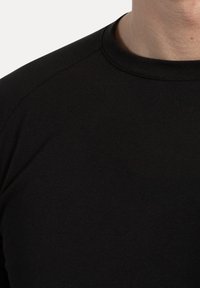 Close-up van een persoon in een eenvoudig zwart T-shirt met ronde hals, waarbij een deel van de nek en schouder zichtbaar is op een witte achtergrond.