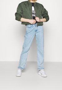 Oliwkowa pikowana kurtka bombka z prążkowym kołnierzem, zestawiona z jasnoniebieskimi prostymi dżinsami i białymi trampkami.