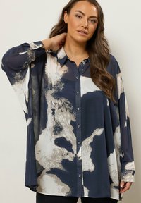 Frau trägt eine lockere, geknöpfte Bluse mit langen Ärmeln und einem abstrakten Muster inMarineblau, Beige und Weiß und posiert mit einer Hand hinter dem Nacken.