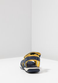 Laivastonsininen sandaali, jossa keltaisia yksityiskohtia, teksturoitu pohja, säädettävät remmit ja yksityiskohtaiset ompeleet kevyessä mallissa.